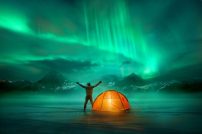 A-man-camping-in-wild-northern-mountains-with-an-illuminated-tent-viewing-a-spectacular-green-northern-lights-aurora-display.-Photo-composition.jpg